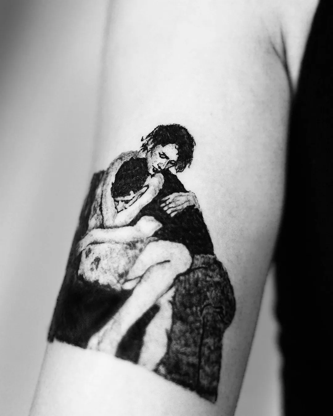 Black and grey blackwork tattoo of two figures embracing, framed on the upper arm